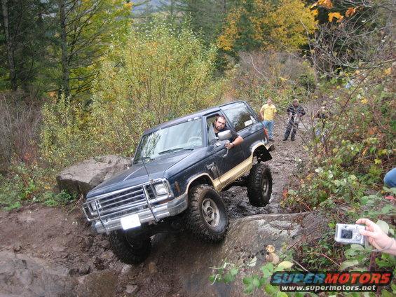 Dark blue Ford Bronco with lifted suspension and off-road tires navigating a rocky trail, driver looking at camera