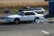 1997 Mercury Grand Marquis - A white sedan performs a burnout on a track, with other vehicles and a cornfield visible in the background.