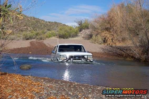 White SUV driving through a shallow river with brush and hills in the background
