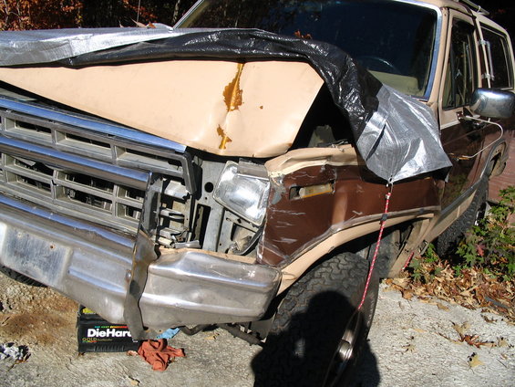 Damaged front end of a tan and brown SUV, covered partially with tarps. A DieHard battery is visible underneath