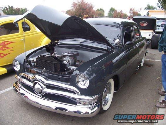Dark gray 1950 Ford sedan, hood open showing engine. Yellow hot rod with flame decals to the left. Other vehicles and people in background