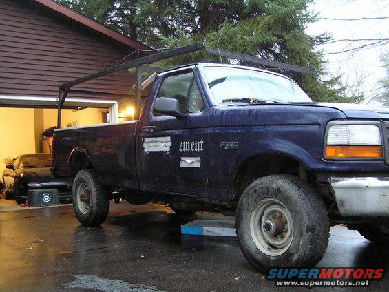 Dark blue Ford F-250 truck with a rack, parked in front of a garage. A black sports car is visible inside the garage