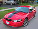 2003 Ford Mustang - This red 2003 Ford Mustang coupe features a prominent black stripe and hood scoop, parked on a residential street.