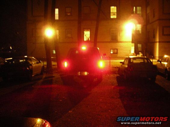 SUV's rear end with brake lights illuminated, parked at night near buildings. Other cars visible