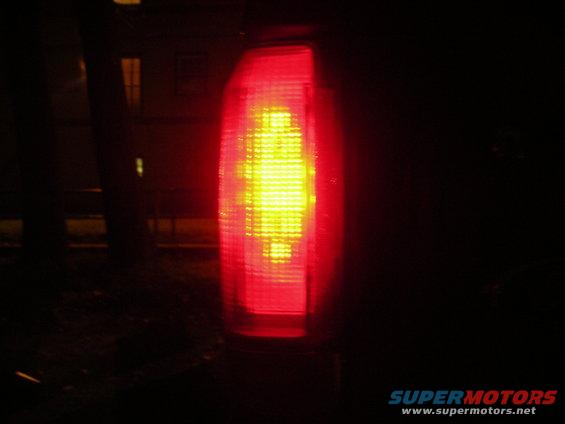 Red taillight with yellow bulb illuminated, dark background, Supermotors.net logo at bottom right