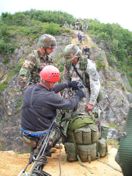 Soldiers in camouflage gear and helmets are rappelling down a rocky, vegetated slope, with ropes and backpacks visible