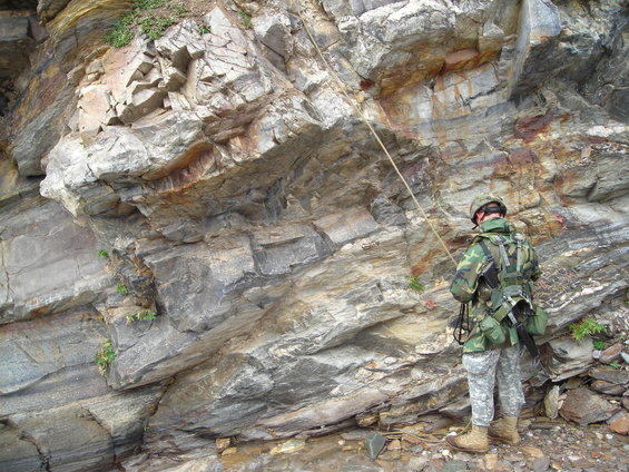 Soldier in camouflage gear with a helmet and backpack rappels down a rocky cliff face with a rope