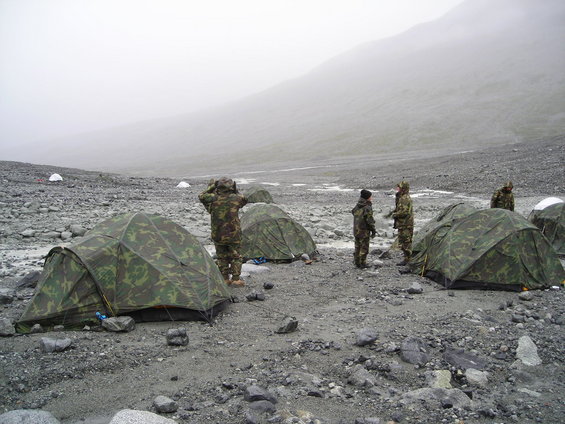 Several camouflage tents are set up on a rocky, barren ground with a foggy mountain in the background. People in camouflage uniforms are standing near the tents