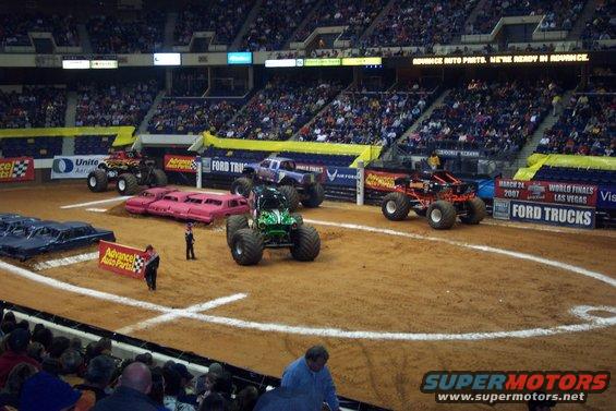 Monster trucks lined up in an arena with spectators in the background. Ford Trucks banner visible