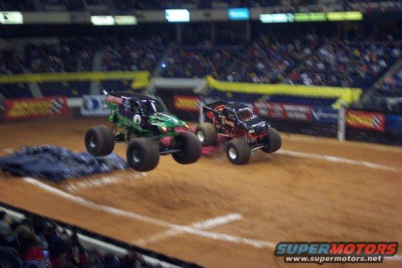 Two monster trucks airborne over a dirt track during a competition