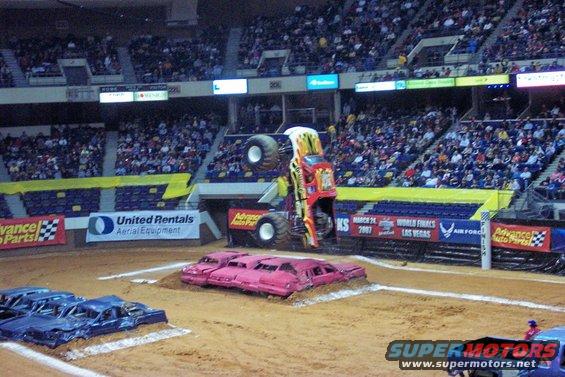Monster truck airborne over crushed pink cars in an arena, with blue cars in foreground