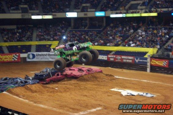 Green monster truck with purple accents airborne over crushed cars