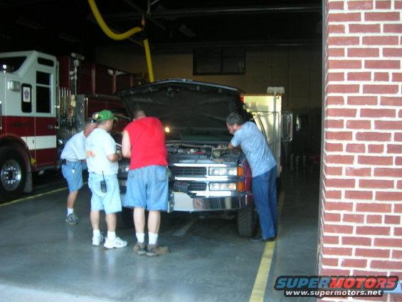 Truck with hood open, people inspecting engine. Red and white fire truck visible to the left