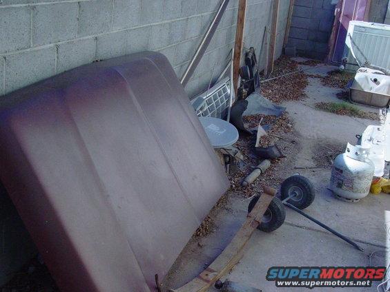 Maroon car hood leaning against a cinder block wall, with various parts and propane tanks scattered around