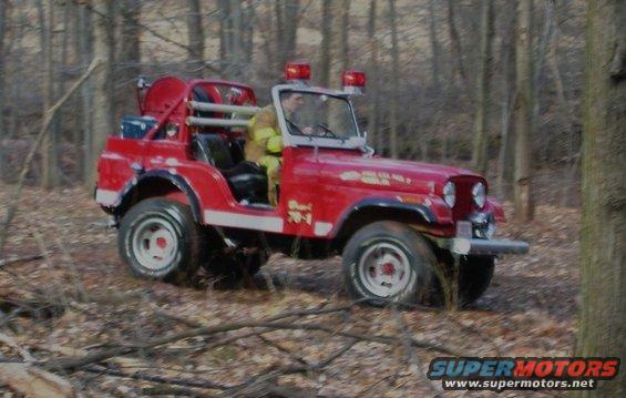 Red Jeep CJ fire truck with firefighter driving through woods. Has a hose reel on back