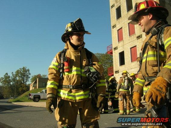 Firefighters in full gear stand outside a brick training facility
