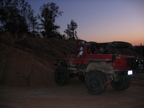 Red off-road SUV with large tires, roll cage, and roof rack, driving on dirt terrain at dusk