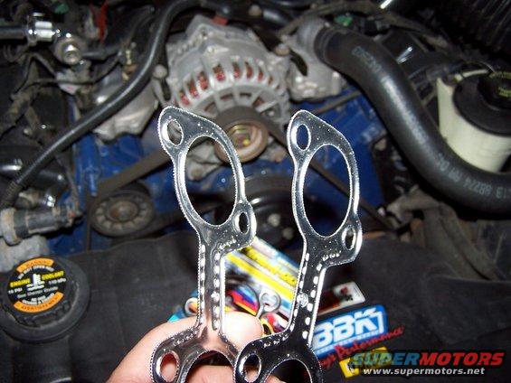 Hands holding exhaust gaskets in front of a car engine bay with alternator and hoses visible