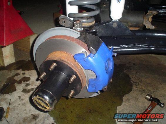 Front wheel brake assembly with blue caliper and rotor, part of a vehicle under repair. Hub cap reads "WARN"