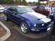 2002 Ford Mustang - A blue Ford Mustang GT with racing stripes is parked in a lot, with other vehicles and people nearby.