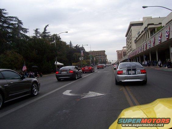 Several Ford Mustangs driving down a street during a parade. One is silver with a license plate that says MUSTANG