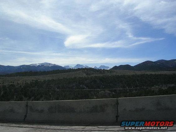 Mountain range under a cloudy sky, with a concrete highway barrier in the foreground. Supermotors.net logo visible