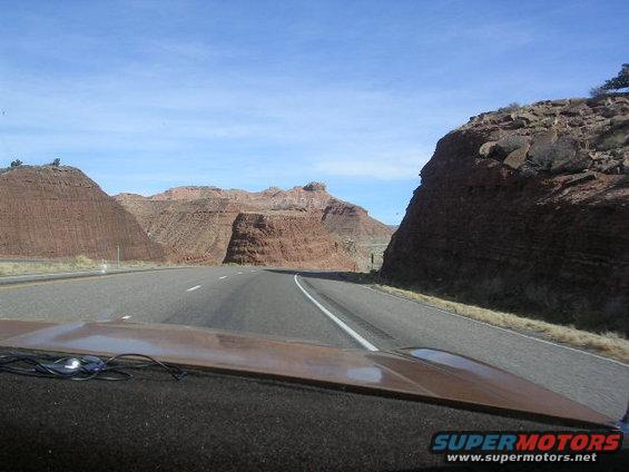 Brown dashboard and hood of a car with wires, viewed from inside, with a highway and red rock formations in the background