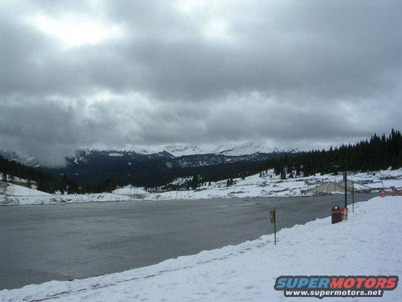 snowy landscape with a frozen lake, pine trees, and mountains under a cloudy sky. A trash can is visible on the right
