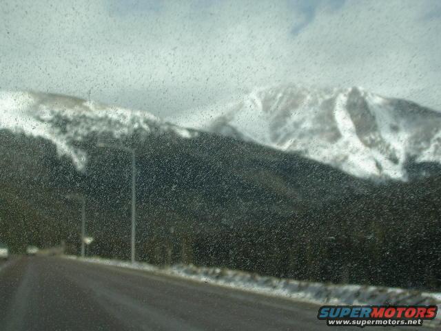 Road leading towards snow-capped mountains under a cloudy sky, seen through a speckled windshield