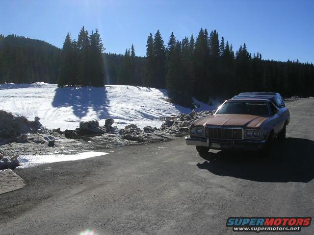 1975 Ford Ranchero with a camper shell parked on asphalt with a snowy hillside and trees in the background