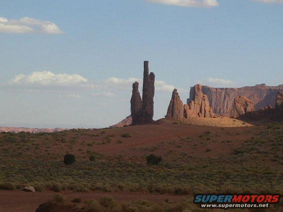 Desert landscape with rock formations and sparse vegetation under a blue sky with clouds. Supermotors.net logo in lower right