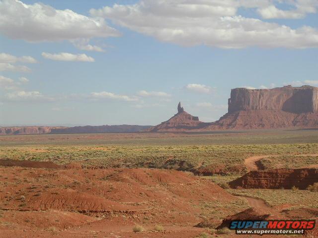 Desert landscape with Monument Valley rock formations and a dirt road, under a cloudy sky. Supermotors.net logo visible