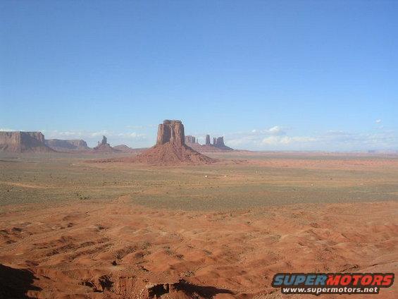 Monument Valley with iconic buttes under a clear blue sky. Foreground shows red desert terrain