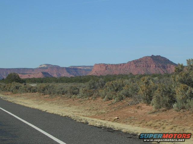 roadside view of desert mesas and scrub brush under a clear blue sky. Supermotors.net logo in corner