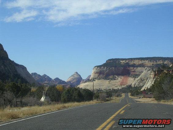 Asphalt road curves through a valley with rocky mountains and sparse trees under a blue sky with clouds