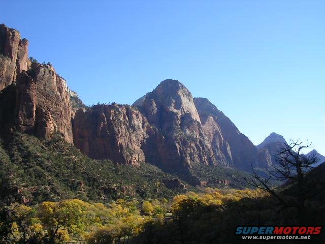 Mountainous landscape with yellow trees under a clear blue sky, Supermotors.net logo in lower right