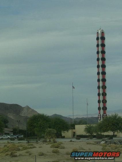 Giant thermometer roadside attraction with flags and parked cars in desert landscape