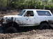 1989 Ford Bronco - This white Ford Bronco is deeply stuck in a muddy off-road area, with trees in the background.