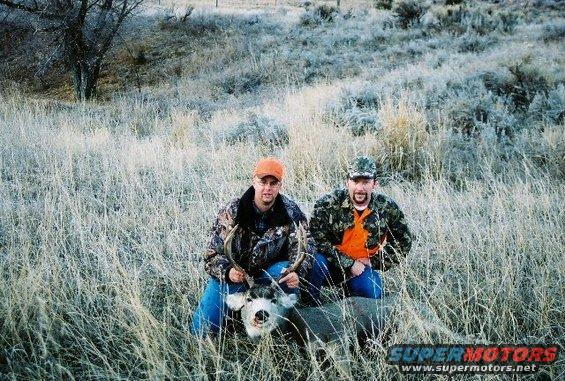 Two men in camouflage kneel in dry grass with a deer carcass between them. One man holds the deer's antlers