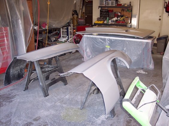 Car fender and bumper cover on sawhorses in a garage, with a hood on a table in the background