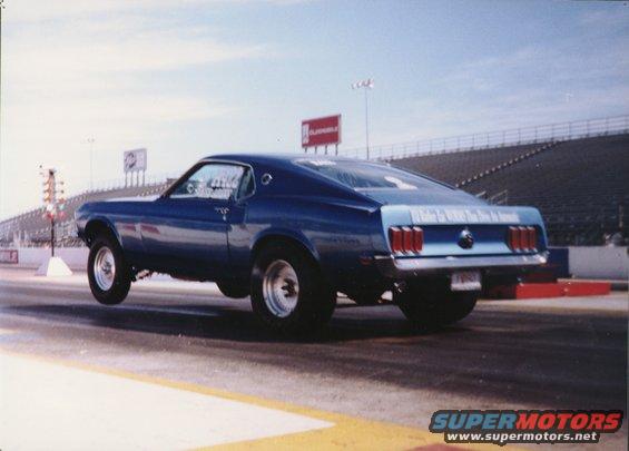 Blue fastback car doing a wheelie on a drag strip. Text on the rear says 'Ford Mustang'