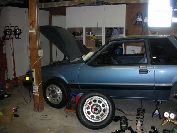 Blue 1990 Ford Mustang coupe side view with hood open and suspension parts on the floor
