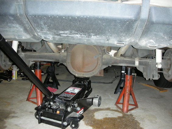 Car's rear axle assembly is supported by jack stands and a floor jack. Fuel tank is visible above