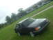1989 Ford Escort - A dark hatchback is parked on a grassy area with a large industrial building in the background.