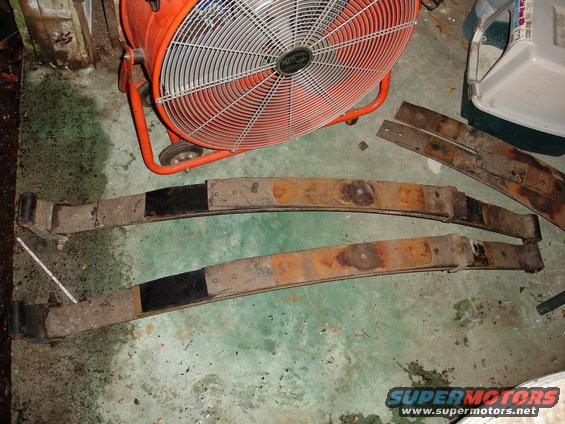 Two rusty leaf springs with black-painted ends sit on a concrete floor next to a large orange fan