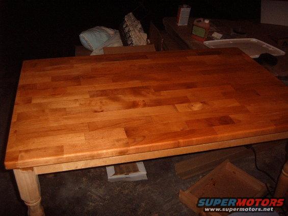 Wooden table with a butcher block top and turned legs. Bottles of finish are on a shelf in the background