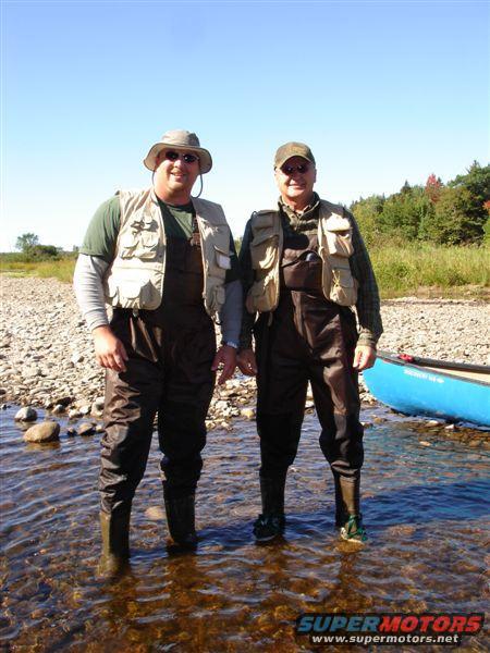 Two guys in waders and fishing vests stand in a shallow river with a blue canoe nearby