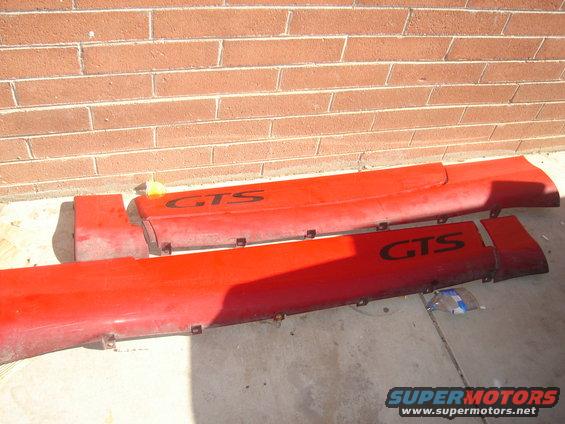 Two red car side skirts with black GTS lettering are laid out on concrete in front of a brick wall