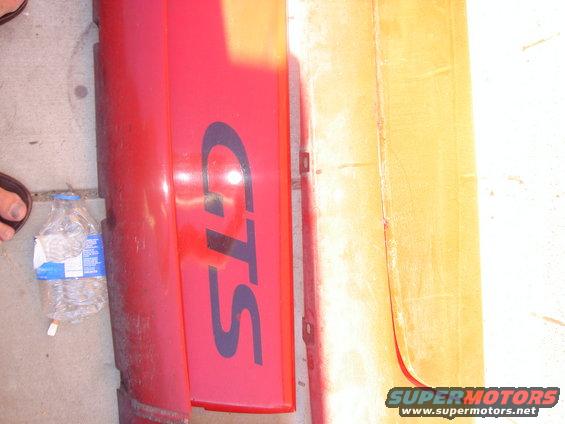 Red car bumper with dark blue GTS lettering, next to a yellowed panel. Water bottle on ground