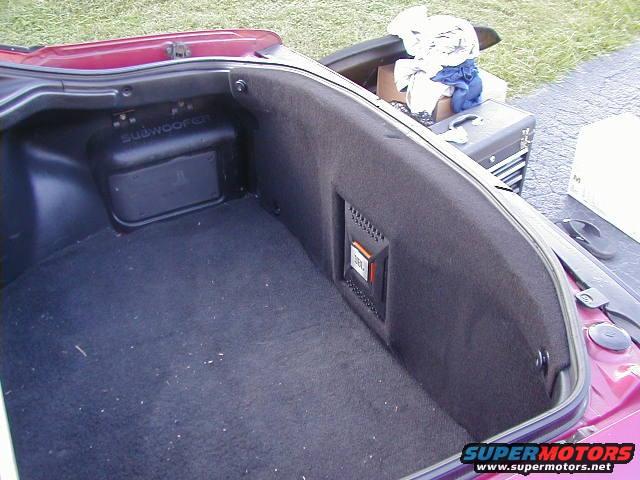Trunk interior showing carpet, subwoofer enclosure, and JBL amplifier. Clothes and a toolbox are visible in the background